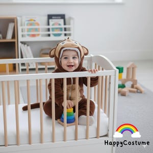 May include: A baby wearing a brown monkey costume sits in a white cot. The costume has a monkey face hood and a long tail. The baby is playing with a colourful stacking toy. The image includes the text "HappyCostume" and a rainbow.