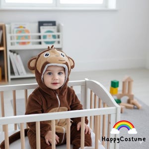 May include: A brown monkey costume with a hood featuring a monkey face, worn by a baby. The costume has a zip closure and a tail. The baby is sitting in a white cot. The text "HappyCostume" is displayed with a rainbow.
