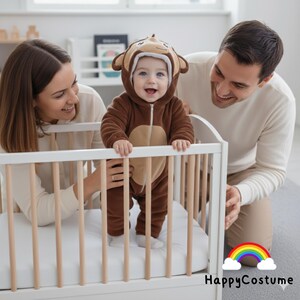 May include: A baby in a brown monkey costume smiles in a white cot. The costume features a monkey face hood and a tan belly. Parents are smiling and looking at the baby. The HappyCostume logo is in the bottom right corner.