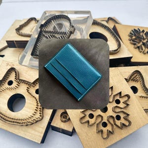 Może przedstawiać: Turkusowy skórzany etui na karty z białymi przeszyciami leży na brązowym kwadracie. Wokół niego znajdują się różne drewniane wykrojniki o różnych kształtach, w tym kota, płatka śniegu i koła zębatego, a także przezroczysty wykrojnik akrylowy.