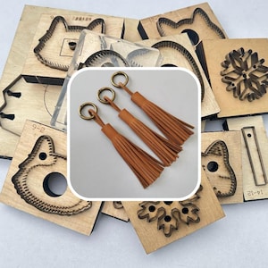 Op de afbeelding: Drie bruine leren kwastjes met goudkleurige sleutelringen. De kwastjes worden getoond voor een verzameling houten stansvormen, waaronder dieren- en sneeuwvlokontwerpen. De stansvormen worden gebruikt voor knutselen.