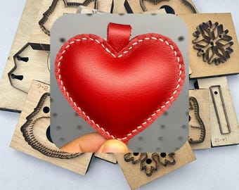 Puede incluir: Un llavero de cuero rojo en forma de corazón con costuras blancas y un lazo. El llavero está rodeado de varios troqueles de madera, incluyendo caras de animales y un copo de nieve.