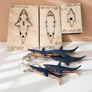 Pode incluir: Chaveiros em forma de tubarão azul marinho e bege, exibidos com modelos de corte de madeira. Os chaveiros são presos a argolas prateadas. Os modelos de madeira têm o texto "GS-237-GD" e "YJ073002".