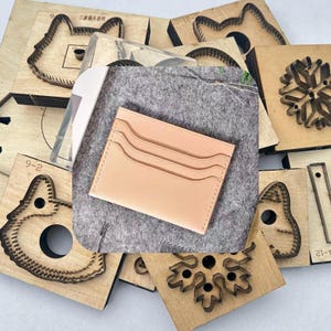 May include: A light tan leather cardholder with three card slots is displayed on a gray felt surface. Surrounding the cardholder are various wooden craft dies in different shapes, including a cat, snowflake, and other geometric designs. The cardholder is a light beige color.