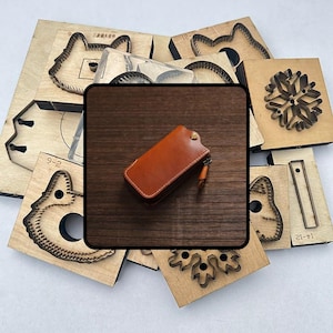 Puede incluir: Un llavero de cuero marrón con cremallera y un pequeño lazo. Alrededor del llavero hay varias formas de troquelado de madera, incluidos diseños de animales y copos de nieve, utilizados para manualidades y trabajos en cuero.