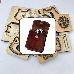 May include: A dark brown leather pouch with a gold-colored snap button closure containing a white USB cable, positioned over a collection of various wooden die-cutting boards with metal blades for crafting shapes like snowflakes and animal heads.