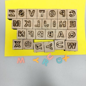 Peut inclure: Tampons de lettres de l'alphabet en bois disposés sur une surface jaune. Les tampons sont disposés en grille, avec des lettres individuelles sculptées. En dessous, des lettres en mousse colorées épellent "M, Y, R, U, G, X".
