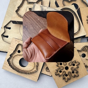 May include: A brown leather wallet with three card slots, held in a hand. The wallet has white stitching around the edges. Surrounding the wallet are various wooden die cut shapes.