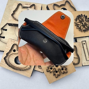 Puede incluir: Estuche de gafas de cuero negro con interior naranja y costuras blancas. El estuche está abierto, revelando un par de gafas con montura negra. El estuche tiene un cierre a presión. Formas de troquelado de madera en el fondo.