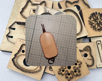 May include: A collection of wooden craft dies in various shapes, including animal faces and a snowflake. A light brown leather key fob with a metal ring and a braided cord is in the center. The dies are arranged on a light-colored surface.