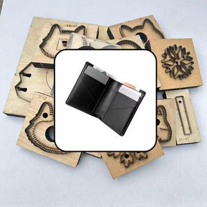 Könnte beinhalten: Ein schwarzes Leder-Bifold-Portemonnaie mit mehreren Kartenfächern und einem Mittelfach, offen dargestellt mit mehreren grauen und weißen Karten. Das Portemonnaie ist vor einem Hintergrund aus verschiedenen hölzernen Stanzformen zentriert.