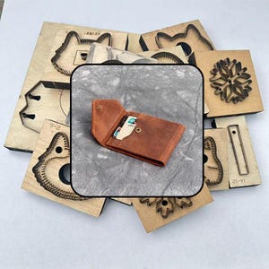 May include: A brown leather wallet with a snap button closure, containing a credit card, positioned over a collection of wooden die-cutting moulds with various shapes including animal outlines and geometric patterns.