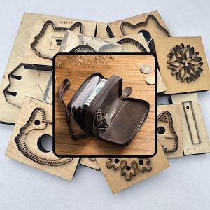 Puede incluir: Un monedero de cuero marrón con cremallera, abierto para revelar dinero en efectivo y llaves, se encuentra sobre una superficie de madera. Alrededor del monedero hay varias matrices de corte de madera en diferentes formas, incluyendo un gato, un copo de nieve y otros diseños geométricos.