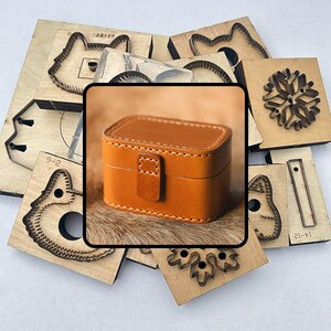 May include: A small, rectangular, tan leather jewelry box with a flap closure. The box is surrounded by various wooden die-cut shapes, including animal and snowflake designs. The box is approximately 3 inches long, 2 inches wide, and 1.5 inches tall.