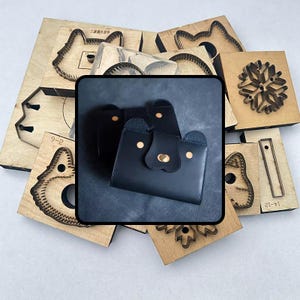 Könnte beinhalten: Eine schwarze Ledergeldbörse mit einem Bärengesicht-Design, mit goldfarbenen Akzenten. Um die Geldbörse herum befinden sich verschiedene Holzstanzformen, darunter Tier- und Schneeflocken-Designs, für Lederarbeiten.