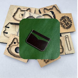 May include: A handmade leather wallet with a dark brown and green design, stitched with white thread, rests on a green leather square. Surrounding the wallet are various wooden craft tools, including die-cut shapes for leatherworking.