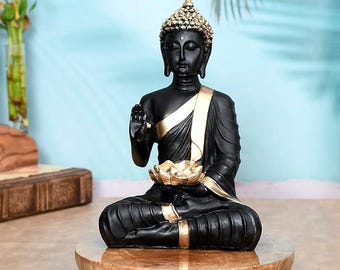 May include: A black Buddha statue in a seated meditation pose, adorned with gold accents on the robe and headpiece. The statue holds a golden lotus flower and sits on a round, wooden stand with small feet. The background is a light blue.