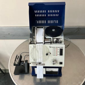 Könnte beinhalten: Eine Greiner vibrograf B500 Maschine mit blauem Oberteil und weißer Front. Die Maschine hat ein Zifferblatt, eine Testtaste und einen Papierausgang. Auf der linken Seite befindet sich ein schwarzer Ständer.