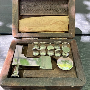 May include: A dark brown wooden box containing a set of silver-colored metal tools. The kit includes a large L-shaped metal component with a threaded adjustment knob, eight small metal attachments with varied shapes, and one circular metal disc.
