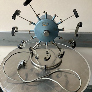 May include: A vintage, light blue, mechanical device with multiple arms and black attachments. The device is circular with a central hub and is mounted on a silver-colored, round table. A power cord extends from the device.