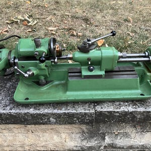 May include: A vintage green metal lathe machine with multiple levers and handles. The machine has a base, a bed, and a headstock. The lathe is designed for shaping metal and other materials. The machine is sitting on a stone surface.
