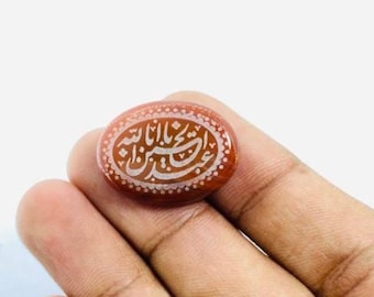 May include: Oval-shaped, reddish-brown gemstone with white Arabic calligraphy. The stone is held in a hand, showcasing the intricate design and color contrast. The calligraphy is the focal point of the stone.