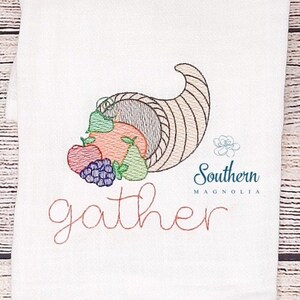 Könnte beinhalten: Besticktes Küchentuch mit einem Füllhorn voller Früchte und dem Text "gather" in einer kursivschriftlichen Schriftart. Der Text "Southern Magnolia" ist ebenfalls rechts unten gestickt.