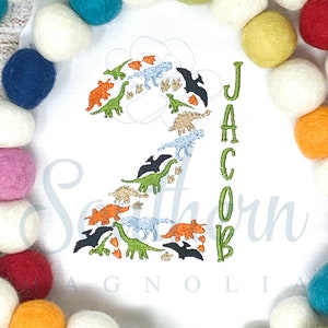 May include: A white onesie with a green number two made of dinosaurs. The name "JACOB" is embroidered in green on the right side of the onesie.