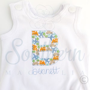 Peut inclure: Combinaison blanche sans manches avec une lettre B brod&eacute;e en bleu et orange. La lettre est remplie de petites images d'avions, de voitures et de trains. Le nom "Bennett" est brod&eacute; sous la lettre.