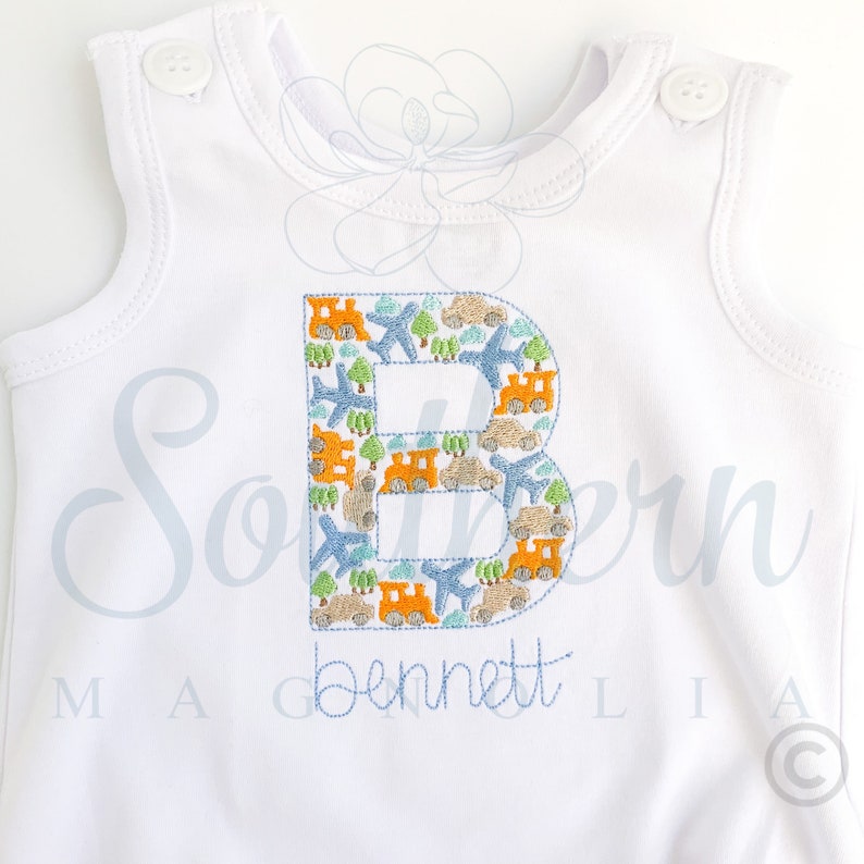 Peut inclure: Combinaison blanche sans manches avec une lettre B brod&eacute;e en bleu et orange. La lettre est remplie de petites images d'avions, de voitures et de trains. Le nom "Bennett" est brod&eacute; sous la lettre.