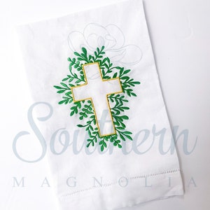 Pode incluir: Toalha de chá de linho branco com folhas bordadas em verde que envolvem uma cruz branca com contorno dourado. O texto "Southern Magnolia" está bordado na parte inferior.