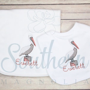 May include: A white burp cloth and bib set with an embroidered pelican and the name "Everett".