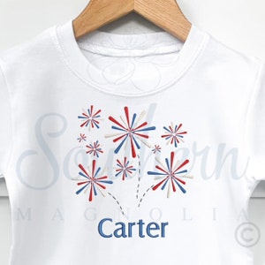 May include: White t-shirt with a red, white, and blue firework design and the name "Carter" embroidered in blue.