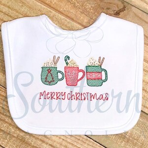 May include: White bib with embroidered red, green, and brown mugs with whipped cream and candy canes. The text "Merry Christmas" is embroidered in red below the mugs.