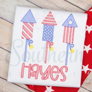 May include: A white t-shirt with a red, white, and blue embroidered design of three fireworks. The text "Hayes" is embroidered in red below the fireworks.