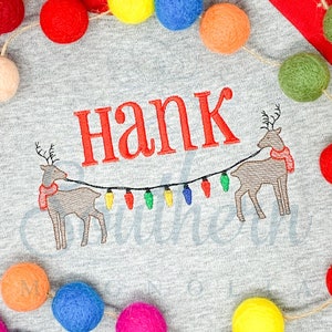 Puede incluir: Sudadera gris con temática navideña con el nombre "Hank" bordado en rojo. Dos renos están bordados en la parte inferior con una cadena de luces.