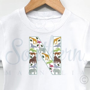 May include: White t-shirt with a large embroidered letter N. The letter is filled with various animal silhouettes, including giraffes, elephants, flamingos, and gorillas.