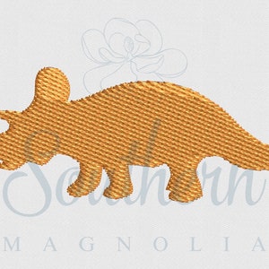Puede incluir: Diseño bordado de un dinosaurio Triceratops dorado. La imagen presenta un patrón detallado y texturizado, con los cuernos, el volante y las patas del dinosaurio claramente definidos. El fondo es una tela clara y texturizada.