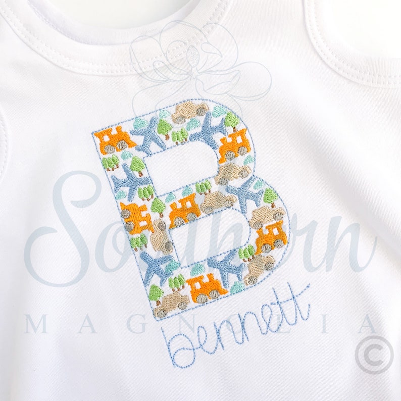 Peut inclure: Une grenouill&egrave;re blanche avec une lettre B brod&eacute;e en bleu. La lettre est remplie d'un motif de petits avions, voitures et maisons bleus, orange et verts. Le nom "Bennett" est brod&eacute; en bleu sous la lettre.