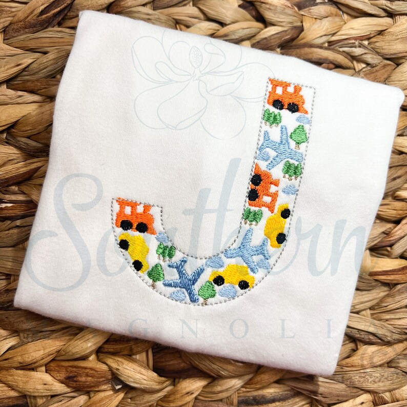 May include: White baby onesie with a blue, yellow, orange and green embroidered letter J. The letter is filled with small images of cars, aeroplanes and trees.