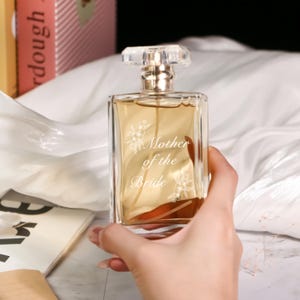 Op de afbeelding: Een heldere, rechthoekige parfumfles met een goudkleurige vloeistof. De fles heeft een heldere, facetgeslepen stop en de woorden "Mother of the Bride" zijn op de voorkant geëtst, samen met bloemmotieven. De fles wordt tegen een witte, zijdeachtige achtergrond gehouden.