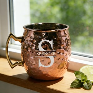 以下が含まれることがあります： ハンマー加工が施された銅製のモスコミュールマグ。ゴールドカラーのハンドルが付いています。「S」の文字と「Spencer」の名前が前面に刻まれています。木製の表面に置かれています。