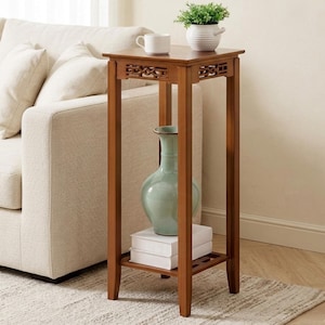 May include: A brown wooden side table with a square top and decorative cutouts. It has a lower shelf and holds a green vase and two white books. A white mug and a small potted plant sit on top. The table is next to a beige sofa. The table is approximately 60 cm tall.