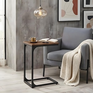 May include: A small side table with a dark brown top and a black metal frame. A gold-coloured mug and a notebook with a pen are on the table. A grey armchair and a cream blanket are in the background.