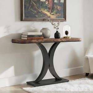 May include: A wooden console table with a dark metal base. The tabletop is a warm brown with a black trim. Decorative items on the table include books, vases, and a glass cloche. The table is against a white wall with a framed painting.