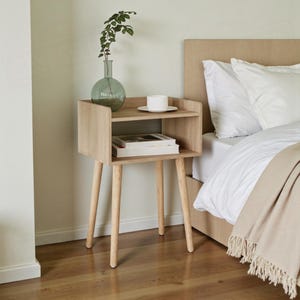 May include: A light oak bedside table with a shelf and three legs. A glass vase with foliage, a white teacup and saucer, and books are on the table. The table is beside a bed with white bedding and a beige blanket.