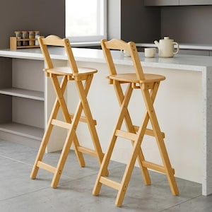 May include: Two light-coloured wooden bar stools with a folding design. The stools have a curved backrest and a round seat. The legs are crossed for stability. The stools are in a kitchen setting.