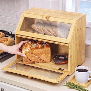 May include: A wooden bread bin with a clear glass door and top, containing a loaf of bread and a chocolate doughnut. The bread bin is open, and a hand is reaching in. Sliced bread is visible inside the top compartment.