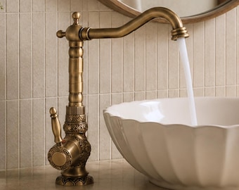 Op de afbeelding: Een messingkleurige metalen kraan met een versierde, getextureerde voet en een gebogen uitloop, die een gestage stroom water in een witte keramische waskom met een geschulpte rand laat lopen. De kraan heeft een enkele hendel en details.