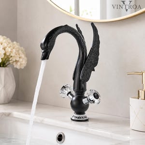 Puede incluir: Un grifo de baño negro mate con forma de cisne, texturas detalladas en las alas y dos manijas de cristal transparente facetado. La base presenta un patrón floral decorativo en blanco y negro, y el agua fluye del pico hacia un lavabo blanco.